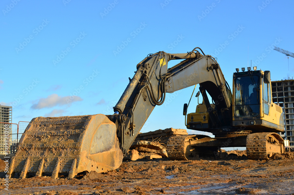 Excavator at a construction site on a background of a construction cranes and building. Backhoe dig the ground for the foundation, laying storm sewer pipes. Installation of water main systems. Excavator at a construction site on a background of a construction cranes and building. Backhoe dig the ground for the foundation, laying storm sewer pipes. Installation of water main systems.