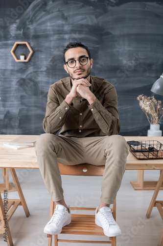 Young restful businessman in casualwear looking at you while sitting on table