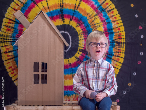 Portrait of funny child in glasses for vision and plaid shirt. boy, with the help of his parents, built cardboard house. kid is pleased with work sitting next to him and enjoys toy house