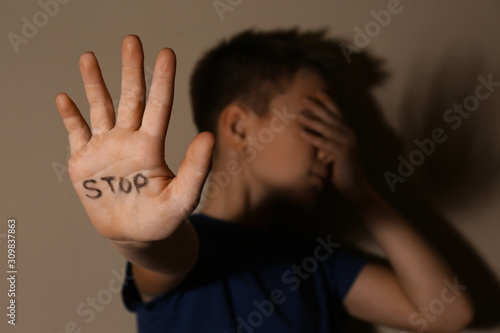 Abused little boy showing palm with word STOP near beige wall, focus on hand. Domestic violence concept