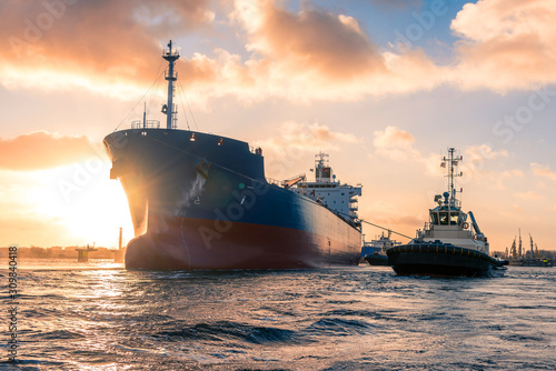 Amazing cloudy sunrise during mooring operations in port. The huge tanker is receiving tugboat assistance to navigate safely to the jetty. Merchant, maritime action background filled with sun rays.