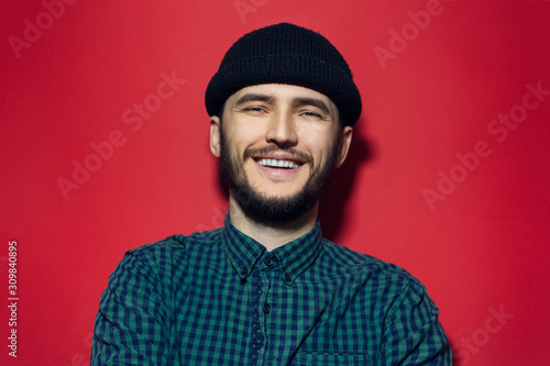 Studio portrait of young smiling man wearing black beanie hat and green plaid shirt on red background. © Lalandrew