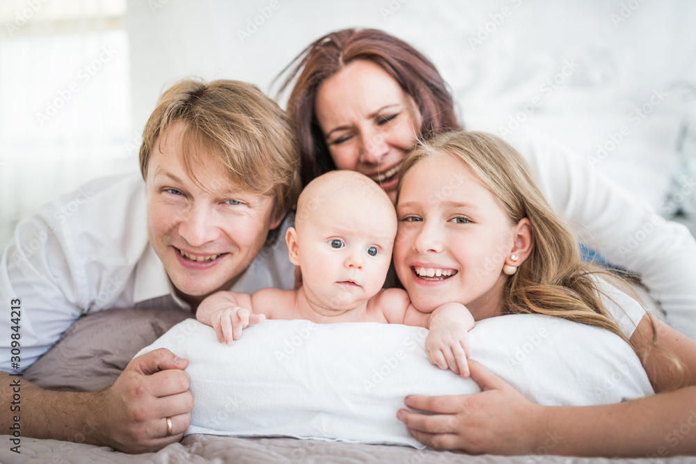Beautiful smiling young family mom dad elder daughter and newborn baby are lying on a large bed in a bright bedroom. Concept of friendly caucasian family