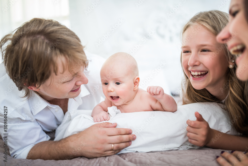 Beautiful smiling young family mom dad elder daughter and newborn baby are lying on a large bed in a bright bedroom. Concept of friendly caucasian family Beautiful smiling young family mom dad elder daughter and newborn baby are lying on a large bed in a bright bedroom. Concept of friendly caucasian family