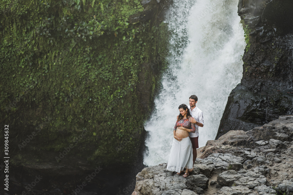 Portrait of happy pregnant spanish couple in love with amazing waterfall view. Happy together, pregnancy travel lifestyle. Maternity wanderlust concept. Portrait of happy pregnant spanish couple in love with amazing waterfall view. Happy together, pregnancy travel lifestyle. Maternity wanderlust concept.