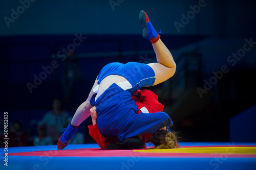 Two woman in blue and red wrestling on a yellow wrestling carpet in the gym..