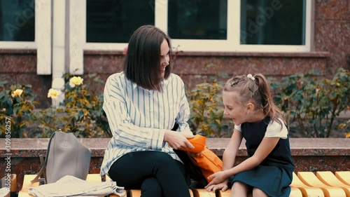 European mother and a little daughter are sitting on the bench. The mother is holding a lunch box of her daughter. The girl is watching with surprise and expecting the meal time. photo