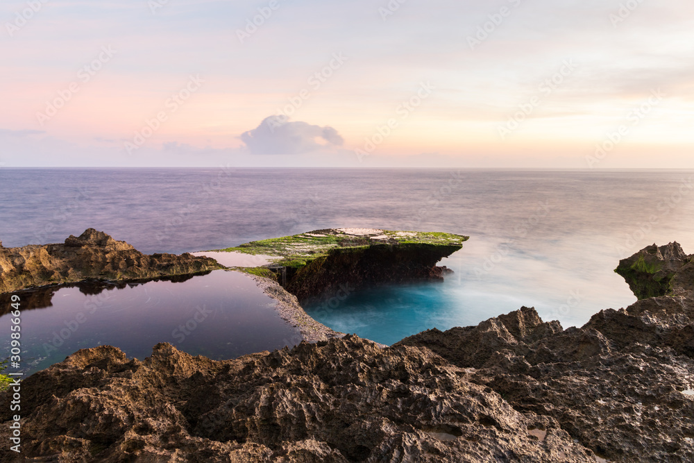 Devil's Tear at sunset, island of Nusa Lembongan, Bali, Indonesia. Rocky shore in foreground. Yellow sky with clouds beyond. 