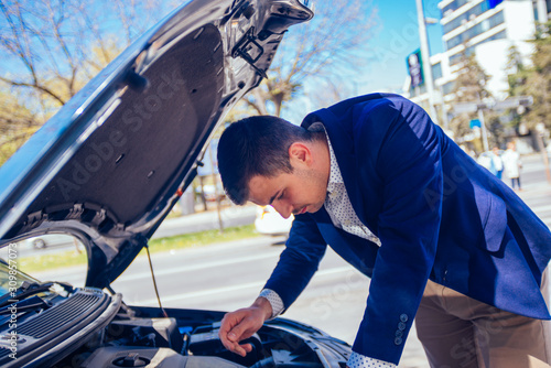 A handsome businessman wearing blue blazer lifting up the hood of his car and checking the oil level on a sunny day parked on a busy city boulevard.