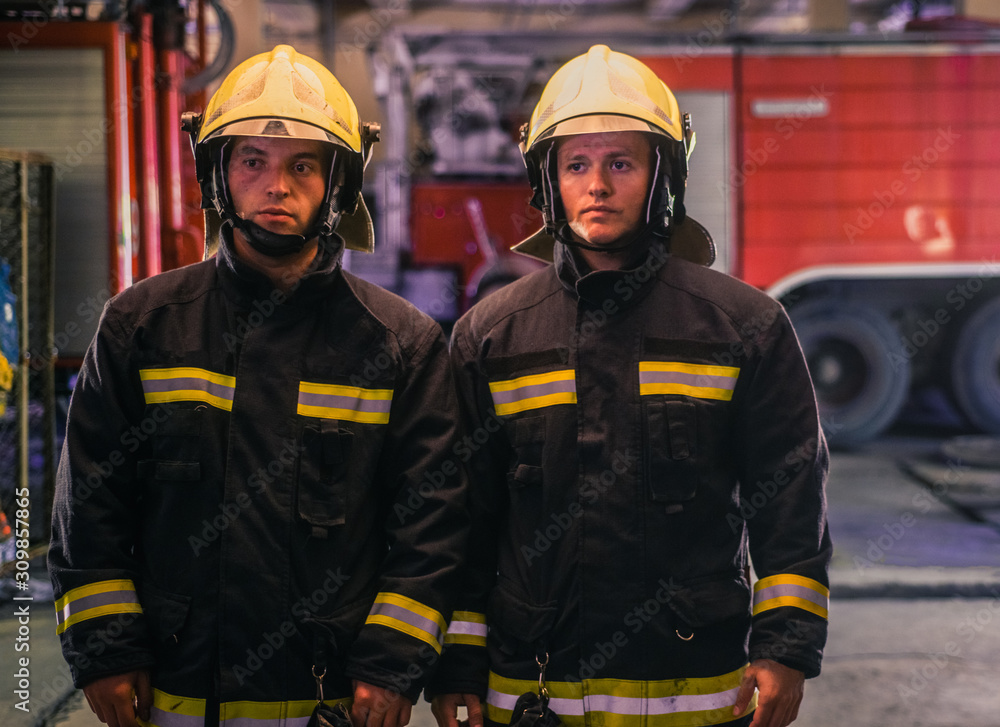 Fototapeta premium Portrait of two young firemen in uniform standing inside the fire station .