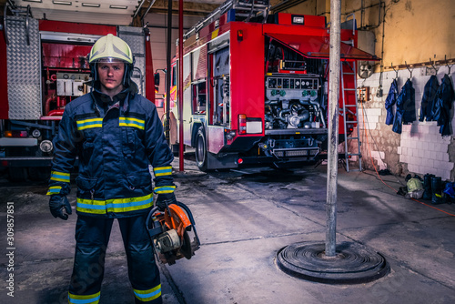 A fireman with uniform and helmet holding a chainsaw with fire truck in the background photo