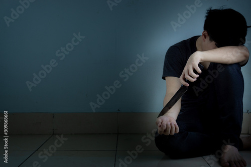sad man hug his knee and cry sitting alone and holding a knife intend to cut his wrist in a dark room. Depression, unhappy, stressed and anxiety disorder concept.
