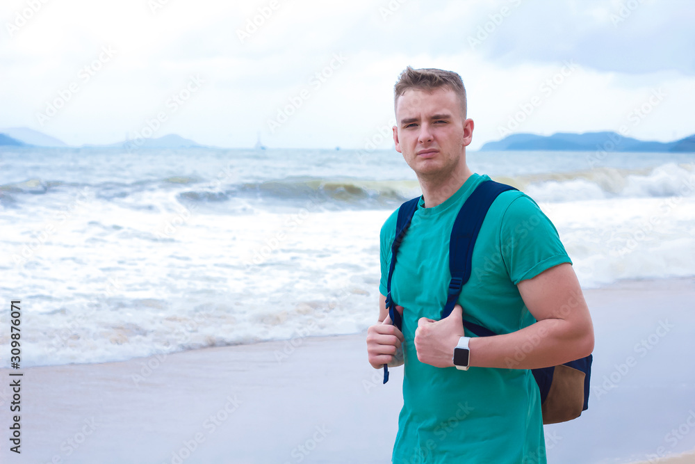 A man travels in a tropical country, walks by the ocean. Displeased, young guy tourist with a backpack on his back. Ustashy handsome man on the background of the sea. Copy Space A man travels in a tropical country, walks by the ocean. Displeased, young guy tourist with a backpack on his back. Ustashy handsome man on the background of the sea. Copy Space