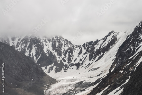 Atmospheric minimalist alpine landscape of big snowy mountain with massive glacier. Low cloud among great rocky mountains. Glacier tongue near snowbound mountainside. Majestic scenery on high altitude
