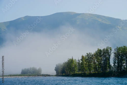Fototapeta Naklejka Na Ścianę i Meble -  Wonderful morning landscape to big mountain on far river bank in fog. Beautiful mist above trees on opposite side of river. Tranquil scenery of pleasant morning freshness in mountains. Inspiring view.