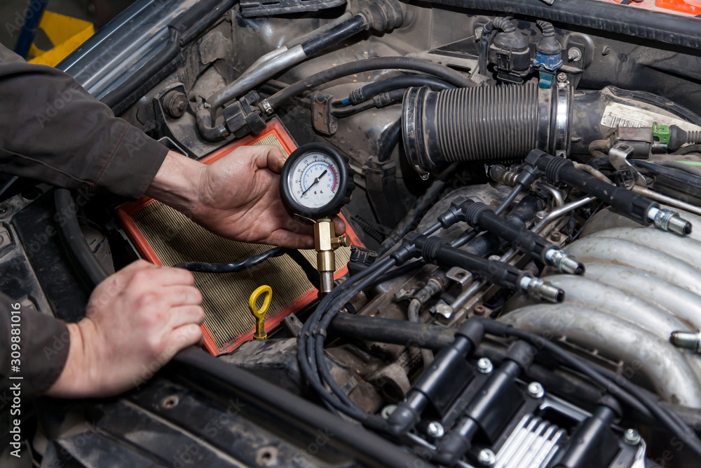 A male mechanic measures the compression in the cylinder of a car engine using a barometer with a scale and an arrow during diagnosis and repair in a workshop for vehicles. Auto service industry.