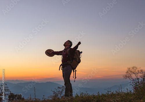 Young man with backpack standing with raised hands on top of mountain at sunset