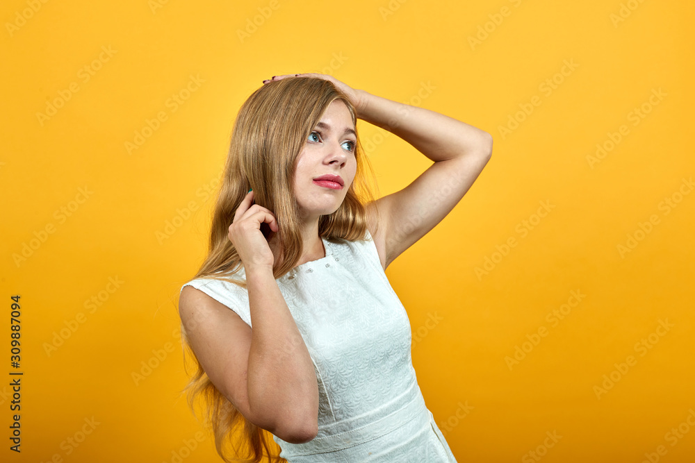 Charming blonde young woman holding hand on head, corrects haircut, looking pretty over isolated orange background wearing white shirt. Lifestyle concept Charming blonde young woman holding hand on head, corrects haircut, looking pretty over isolated orange background wearing white shirt. Lifestyle concept