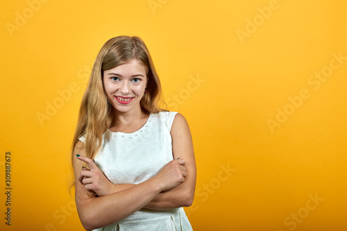 Charming blonde young woman keeping hands crossed, hugging herself, smiling, looking at camera over isolated orange background wearing white shirt. Lifestyle concept