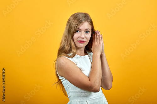 Sleepy blonde young woman keeping hands together, doing sleep gesture, looking at camera over isolated orange background wearing white shirt. Lifestyle concept