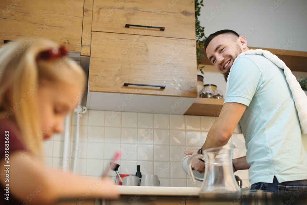 Young bearded father washing dishes and smiling while looking to her father which is drawing in the kitchen while mother is working. Young bearded father washing dishes and smiling while looking to her father which is drawing in the kitchen while mother is working.