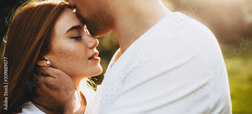 Side view of a charming young couple while man is kissing on forehead her red haired girlfriend which is with closed eyes against sunset.