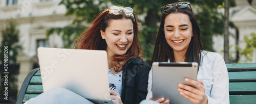 Two charming young girlfriend laughing while looking to a tablet screen sitting on the bench after lessons in a park.