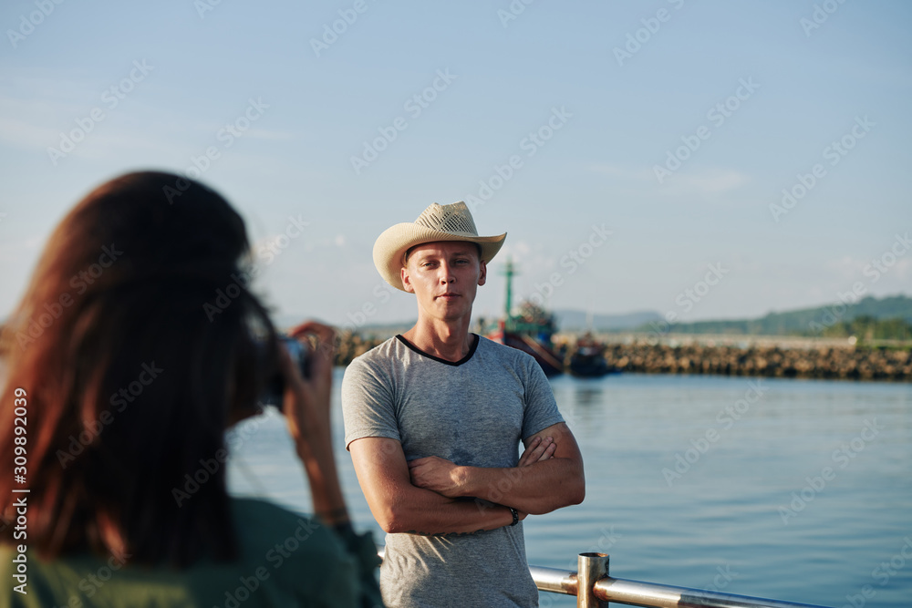 Young woman taking selfie of husband posing in hat against the small bay