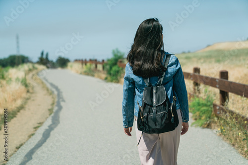 back view of asian woman traveler happy relaxing in grassland outdoor under blue sky with sunlight. young girl backpacker walking on road path way between mountain forest tree and wood vintage fence