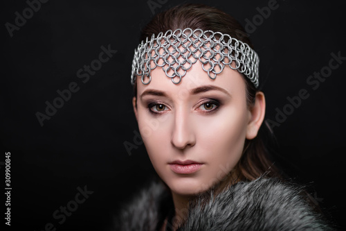 beauty portrait of a girl with a chain mail strip on her head and fur on her shoulders. Studio photo on a black background. Model with clear skin.