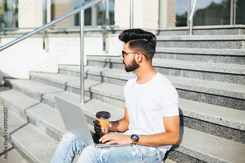 Handsome young indian man working on laptop while sitting outdoors on the stairs.