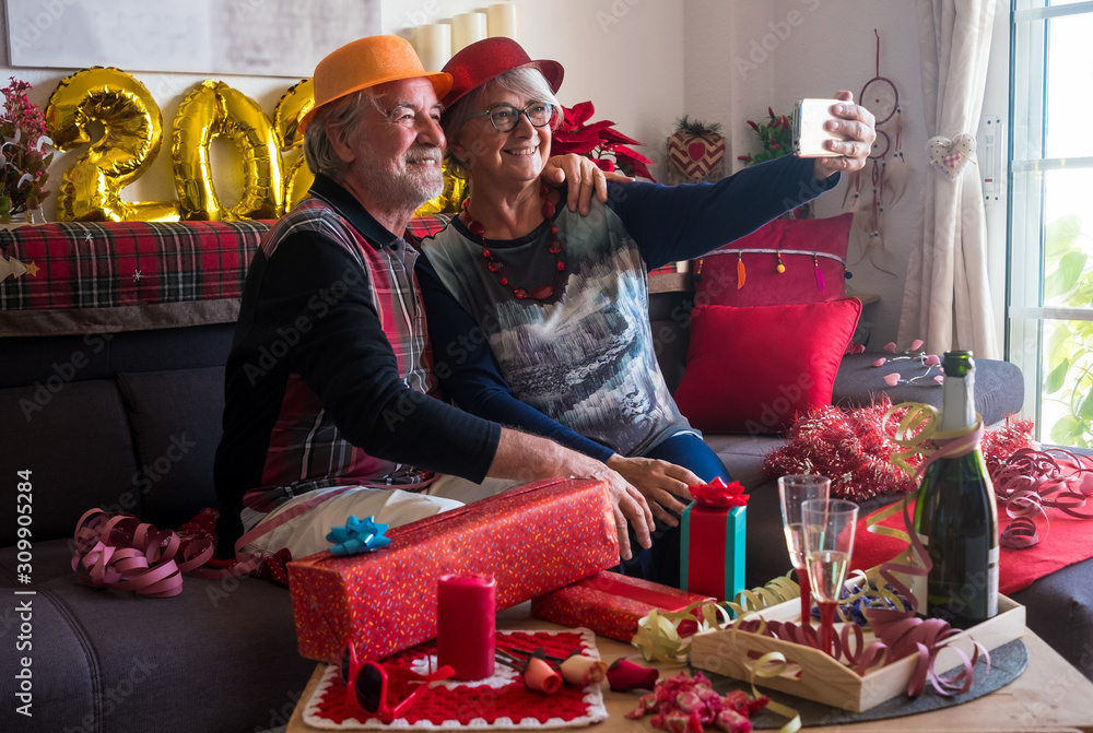 Cheerful couple of senior people with hat sitting on the sofa and looking at mobile phone for a selfie. Toasting with a sparkling wine. Two people Cheerful couple of senior people with hat sitting on the sofa and looking at mobile phone for a selfie. Toasting with a sparkling wine. Two people