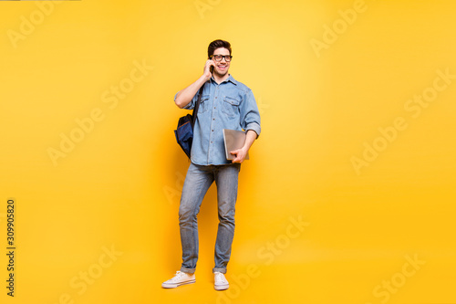 Full length body size photo of cheerful positive toothy beaming man with laptop in hand speaking by phone in white sneakers isolated vibrant color background