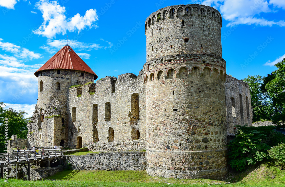 Cesis, Latvia - Cesis castle, stone walls and towers, a bright brown roof, against a background of green grass and a blue sky with clouds, a small wooden bridge with people. 