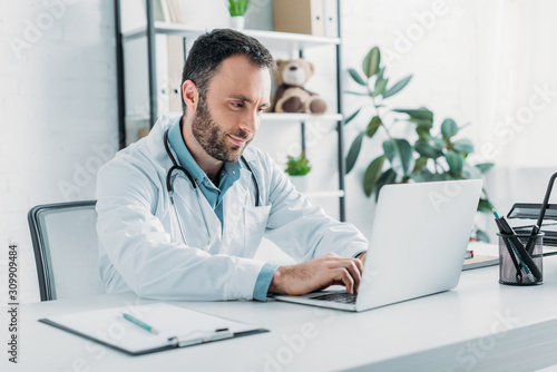 positive doctor sitting at workplace and using laptop photo