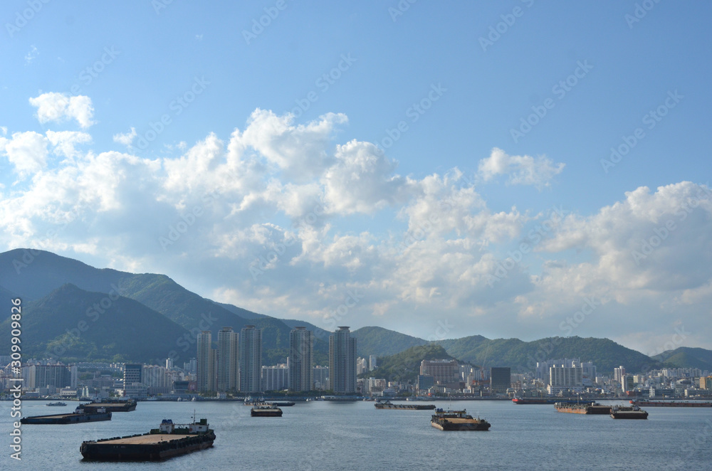 Apartment blocks line the Port of Busan in South Korea, with forest-covered mountains behind them. Barges are waiting to collect cargo. The sky is blue with white clouds.