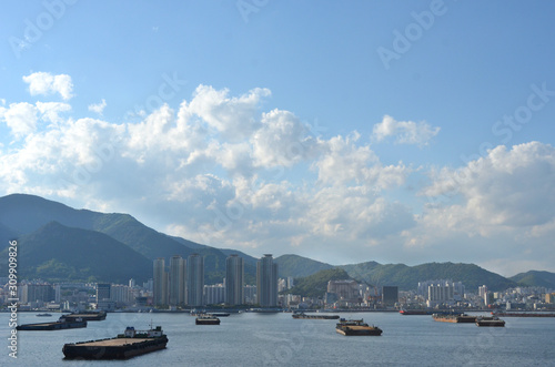 Apartment blocks line the Port of Busan in South Korea, with forest-covered mountains behind them. Barges are waiting to collect cargo. The sky is blue with white clouds. © Anne