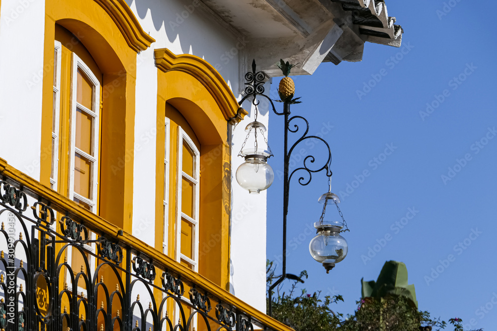 Close up of an artful colonial house corner with two yellow framed high glass doors, metall balustrade and a lantern in sunshine, historic town Paraty, Brazil