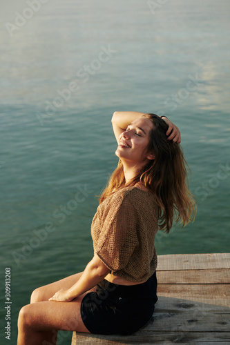 Smiling beautiful young woman sitting on wooden pier, touching her hair and enjoying sunset sun rays © DragonImages
