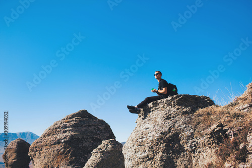 A guy with a backpack sits on a rock in the mountains, drink tea and enjoying view of nature. Nature background. Journey. A trip to the mountains.