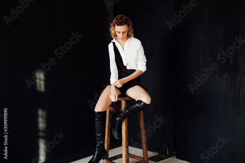 Beautiful fashionable blonde woman in a white blouse and black shorts with boots on a black background