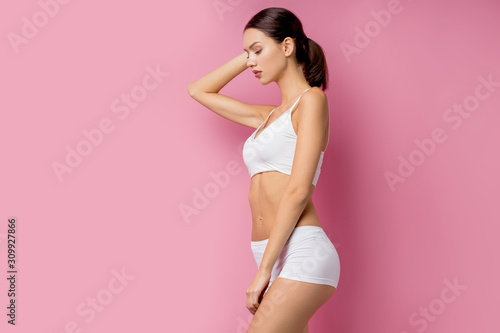 portrait of beautiful slim female in white lingerie isolated over pink background, young brunette with sportive body stand looking down and posing. portrait