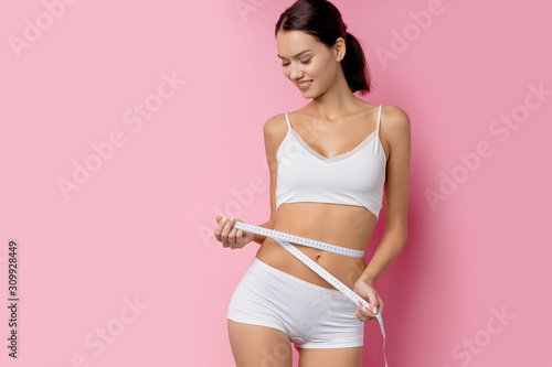 slim woman measuring her body with white metric tape measure after a diet, isolated over pink background photo