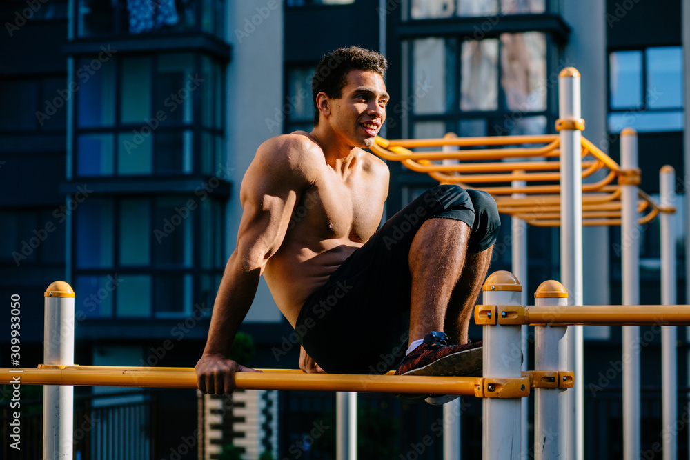 Portrait of athlete young mixed race man resting after exercises on horizontal crossbar at sportsground. Handsome muscular shirtless male training hard, sportsman working out outside.