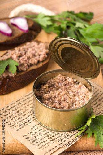 Canned fish, in an open tin, two pieces of dark bread, with onions and parsley, on a wooden cutting board, brown background, close up, vertical