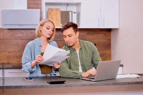 couple checking analyzing utilities bills sitting together at kitchen table, husband and beautiful wife reading bank loan documents with laptop, family managing finances planning expenses together