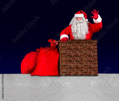 Fotografia Cheerful Santa Claus peek out from a chimney on snowy roof with bags of Christmas gifts and waving with his hand
