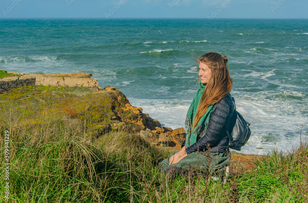 Fototapeta premium Woman dreamily looks at the Atlantic ocean and smiles, sits on the edge of a rocky shore, the wind blows hair, makes waves. Copy space.