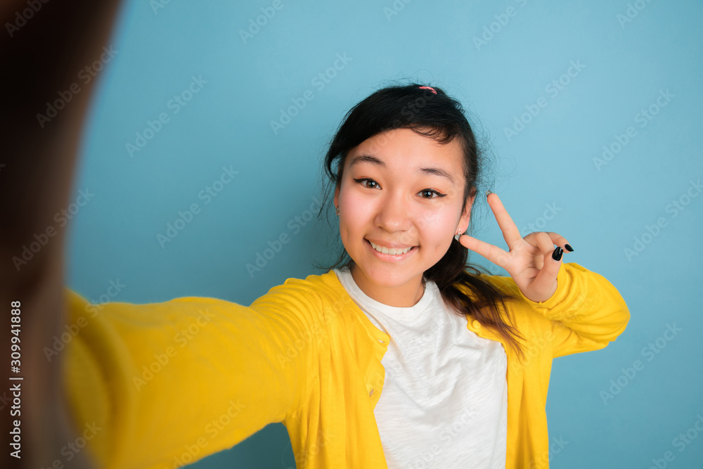 Asian teenager's portrait isolated on blue studio background. Beautiful female brunette model with long hair. Concept of human emotions, facial expression, sales, ad. Making selfie in casual clothes. Asian teenager's portrait isolated on blue studio background. Beautiful female brunette model with long hair. Concept of human emotions, facial expression, sales, ad. Making selfie in casual clothes.