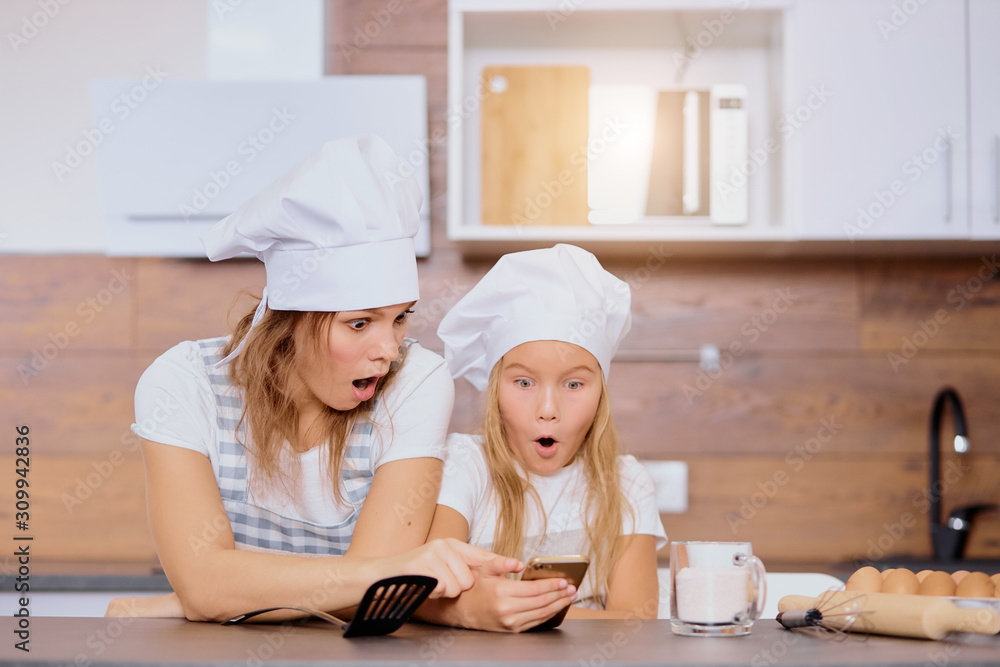 woman and kid girl looking for recipe, stay in shock, before baking cakes, wearing aprons and caps in kitchen woman and kid girl looking for recipe, stay in shock, before baking cakes, wearing aprons and caps in kitchen